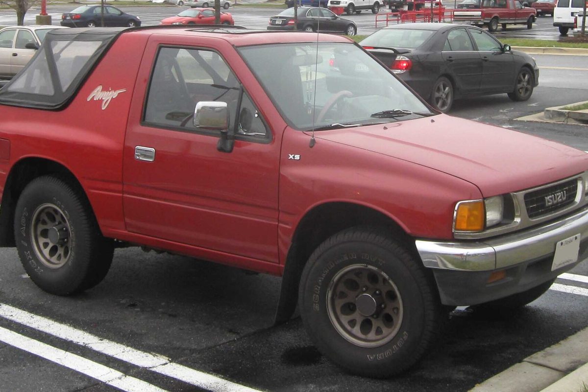 A red Isuzu Amigo XS with a black soft top over the rear, a chrome front bumper and steel wheels, parked in a lot