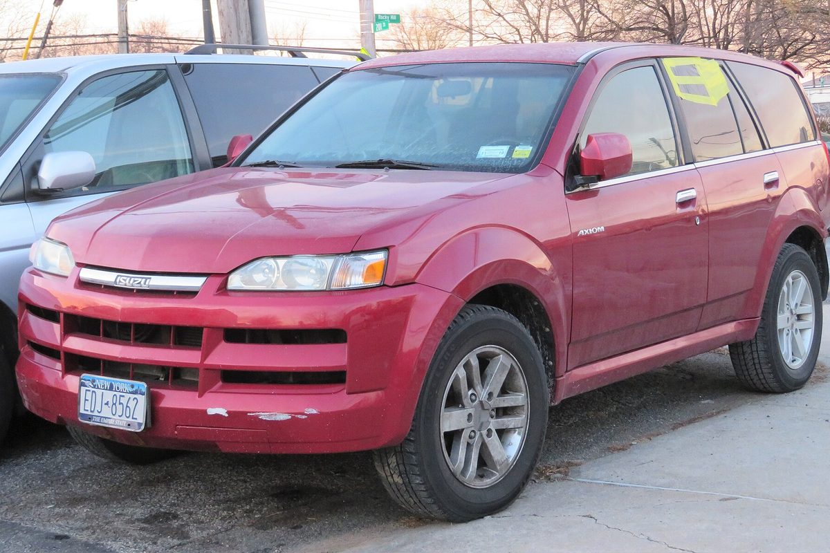A red 2002 Isuzu Axiom SUV with chipped paint on the front bumper and a sign in the windshield, parked by a curb