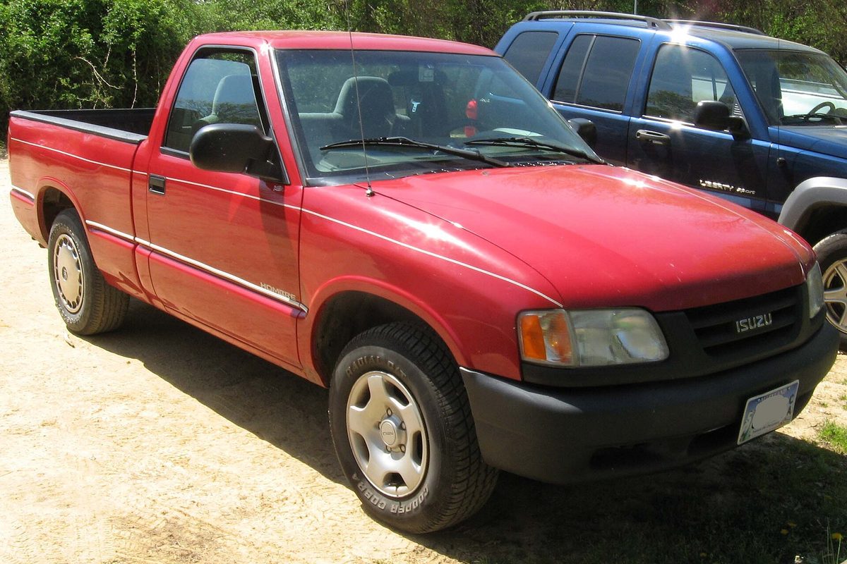 A red regular-cab Isuzu Hombre pickup with black bumpers and steel wheels, parked on dirt in front of green bushes
