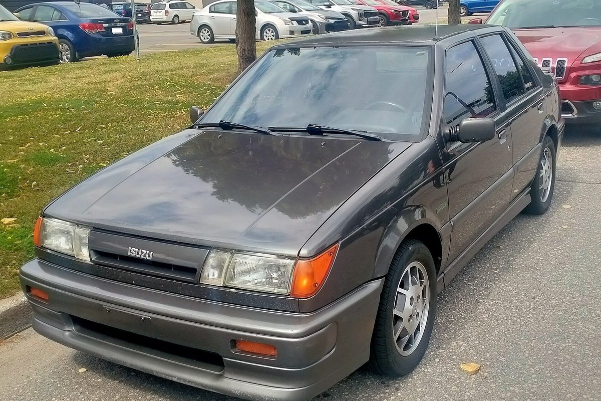 A dark gray Isuzu I-Mark hatchback with alloy wheels, parked on a street beside a grassy strip