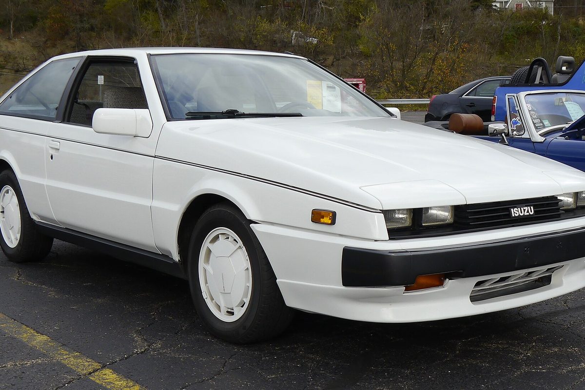A white first-generation Isuzu Impulse hatchback coupe with white wheel covers, parked in a lot with other cars