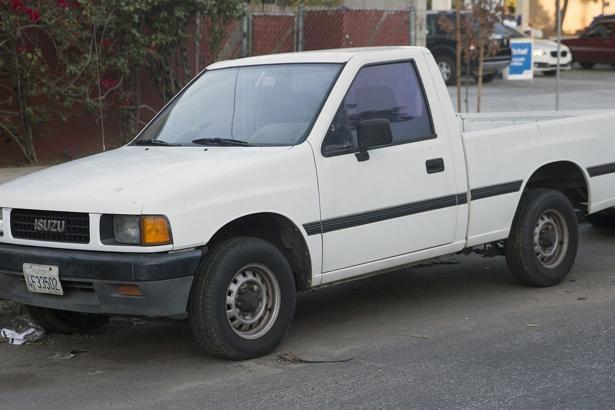 A white regular-cab Isuzu Pickup with a black side stripe and steel wheels, parked at a curb by a fence