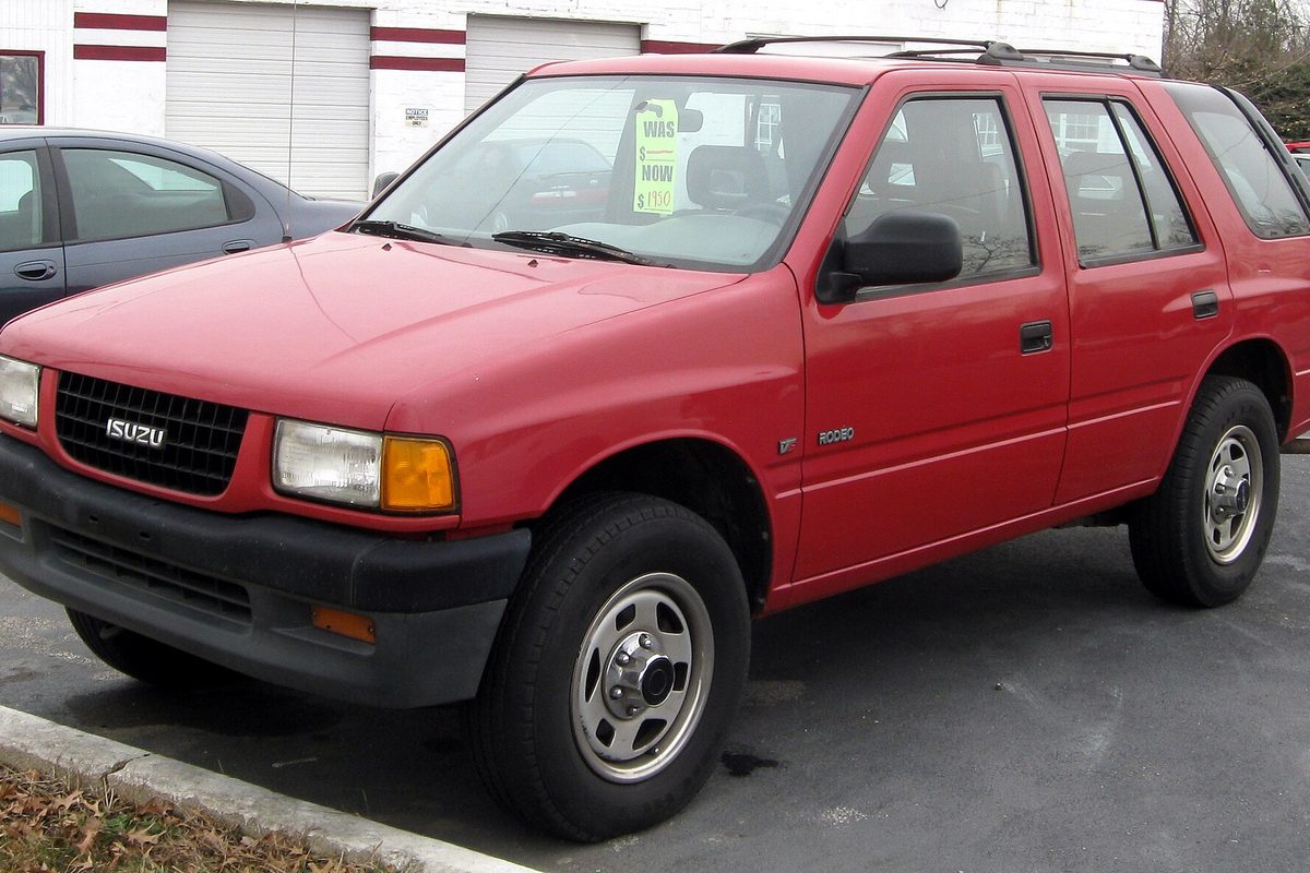 A red 1995 Isuzu Rodeo four-door SUV with steel wheels and a sale sign in the windshield, parked at a curb