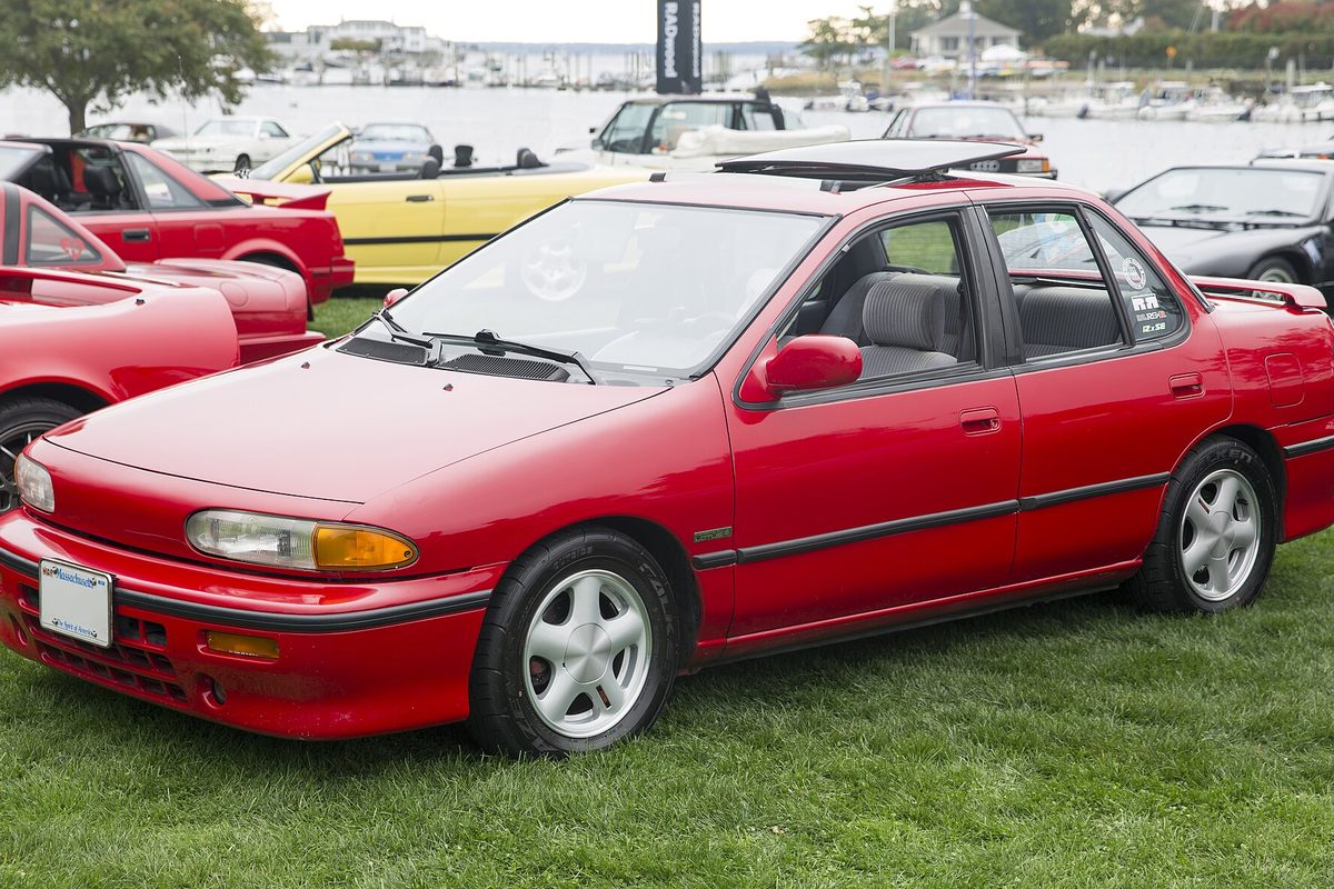 A red Isuzu Stylus sedan with a sunroof and silver alloy wheels, parked on grass at a car show by the water