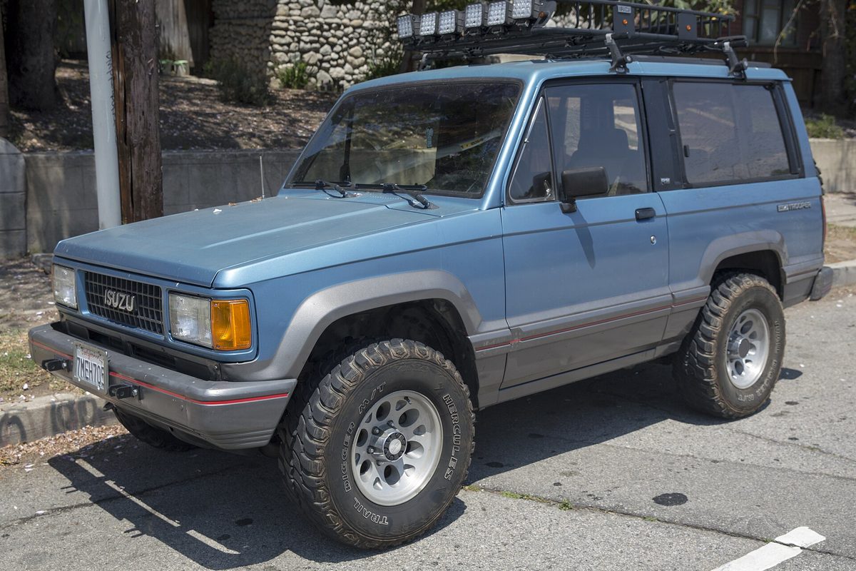 A light blue 1989 Isuzu Trooper II two-door with gray lower trim, big off-road tires and a light bar on the roof rack, parked on a street