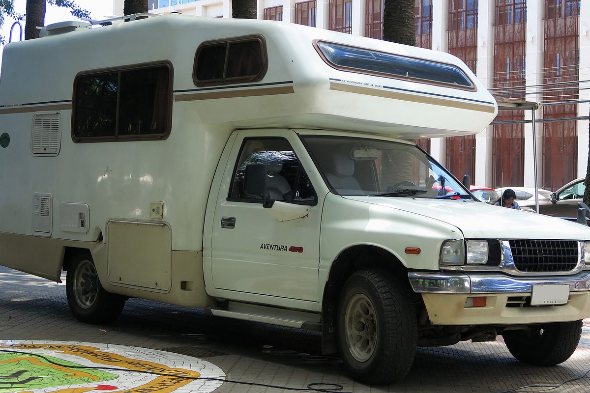 A white 1992 Isuzu pickup badged Rodeo 2.8d, the name it carried in Chile, with a camper body built over its bed, parked on a paved plaza
