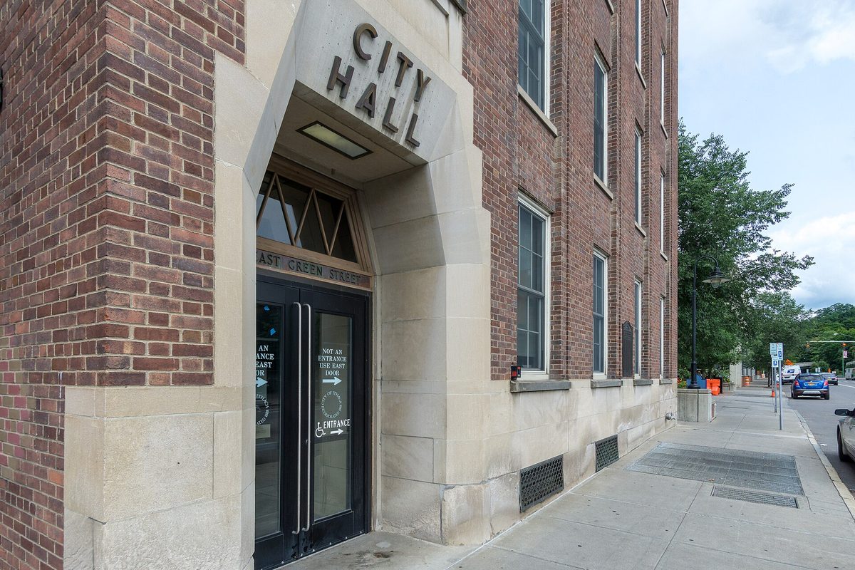 The stone entrance of Ithaca City Hall on East Green Street, with CITY HALL carved above glass doors and a red brick facade beside it