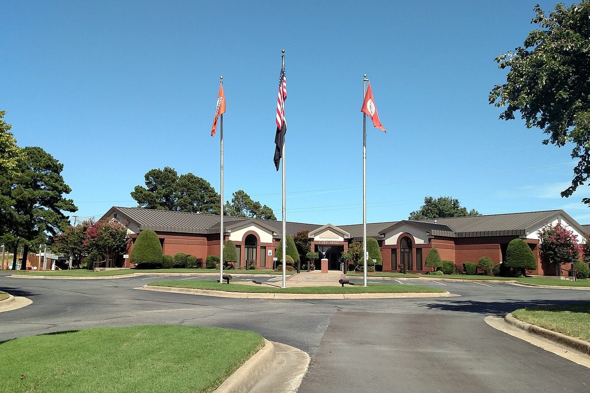 Jacksonville City Hall, a low red-brick building behind a circle drive with three flagpoles flying the US, Arkansas and city flags under a clear blue sky