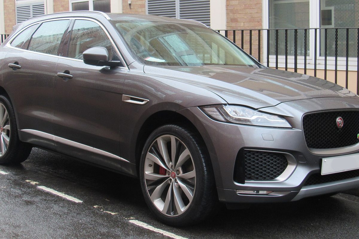 A dark grey Jaguar F-Pace SUV with big alloy wheels and red brake calipers, parked on a wet street beside a black iron railing
