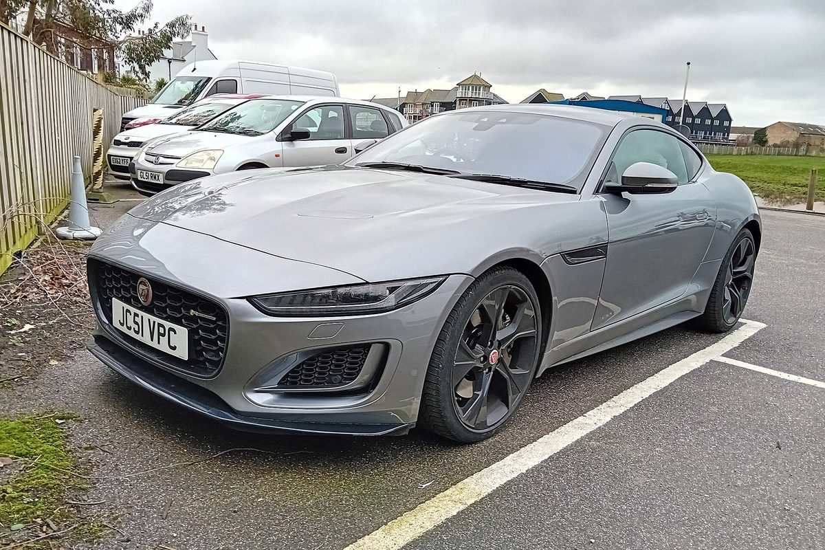 A grey restyled Jaguar F-Type coupe with black wheels, parked in a gravel lot with white cars and a van behind it