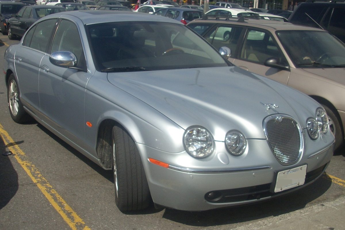 A silver Jaguar S-Type sedan with a mesh grille and four round headlights, parked in a lot marked with yellow lines