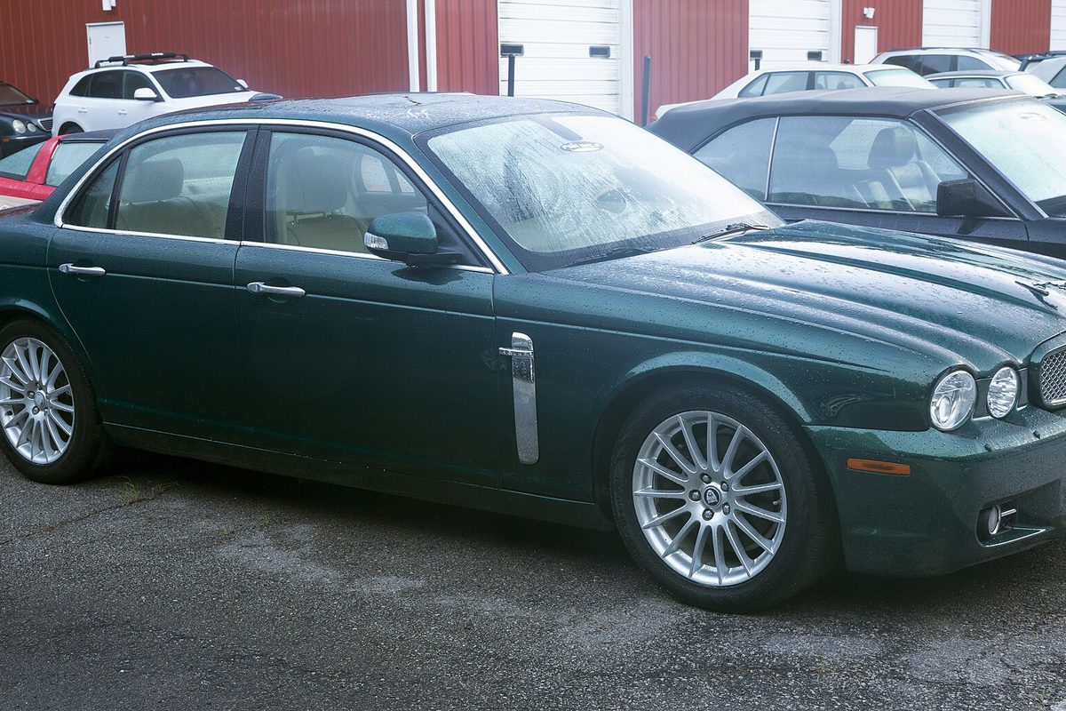 A dark green Jaguar XJ8 sedan, wet with rain, parked on an asphalt lot in front of a red building