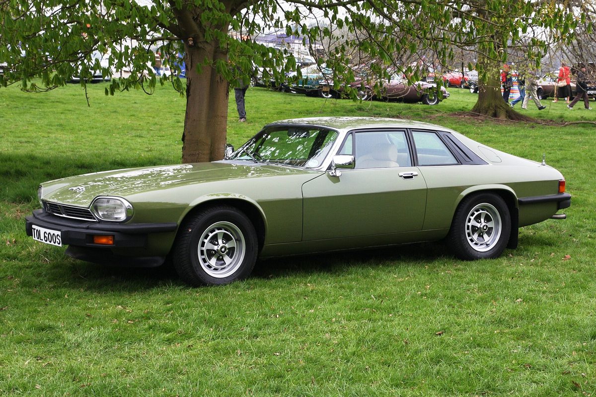 An olive-green early Jaguar XJ-S coupe with silver alloy wheels parked on grass under a tree at an outdoor car show
