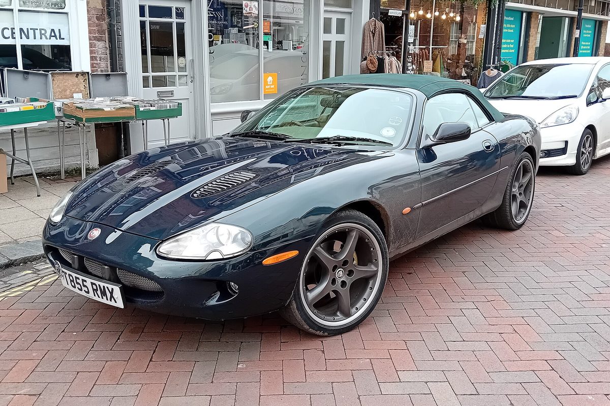 A dark green Jaguar XKR convertible with its soft top up, parked on a brick-paved shopping street