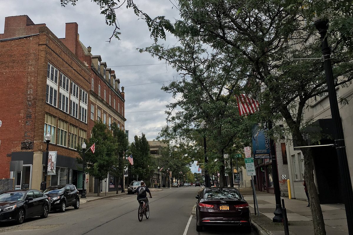 East Third Street in downtown Jamestown, with tall brick buildings on the left, parked cars, street trees and a cyclist riding down the lane