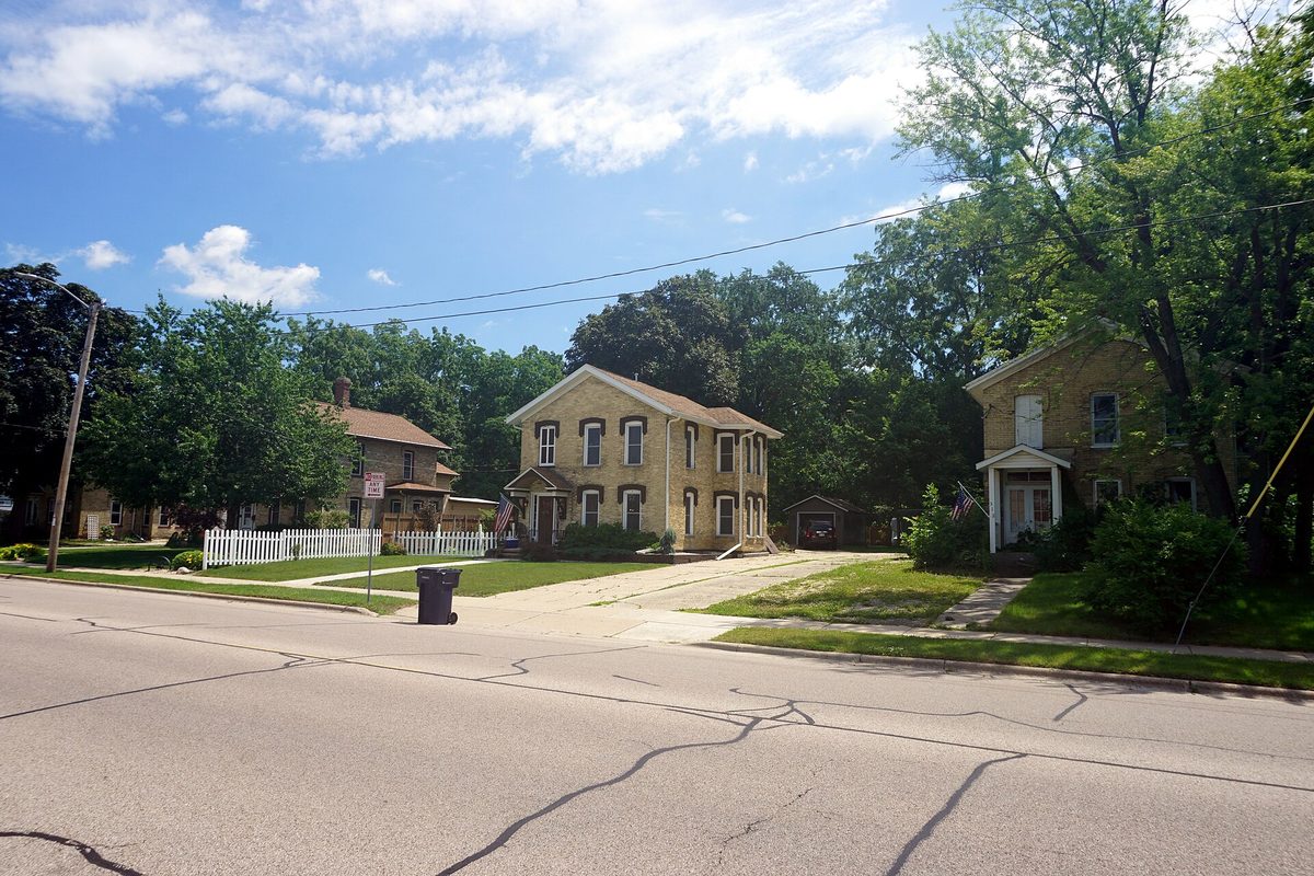 Two cream-colored brick houses with dark shutters on a quiet, tree-lined residential street in Janesville's Conrad Cottages Historic District