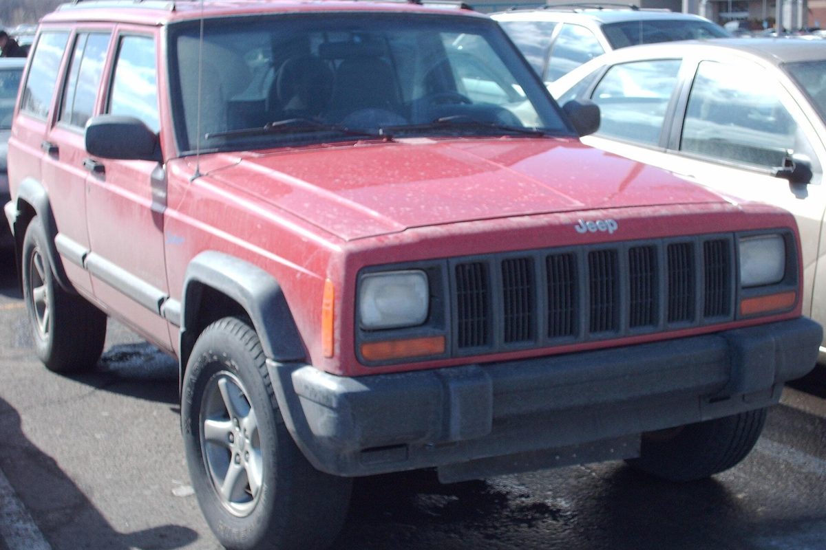 A faded red four-door Jeep Cherokee XJ with black bumpers and gray fender flares, dusty and parked in a lot