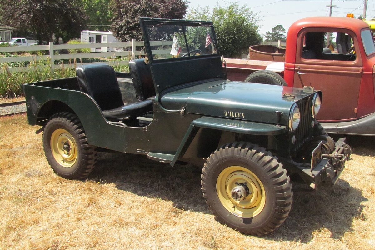 A dark green 1940s Willys CJ-2A with an open top, upright windshield and cream wheels, parked on dry grass in front of an old red truck