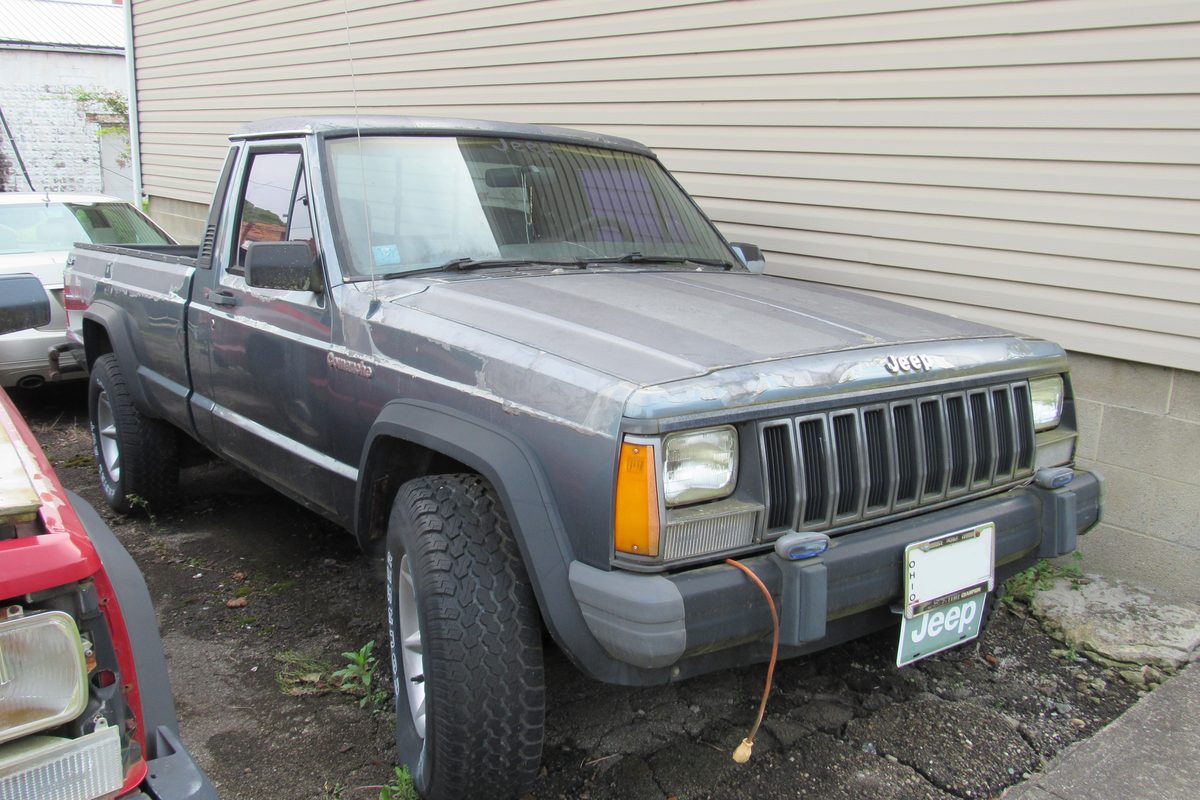 A gray Jeep Comanche regular-cab pickup with darker gray lower trim and worn paint, parked on gravel beside a house with beige siding