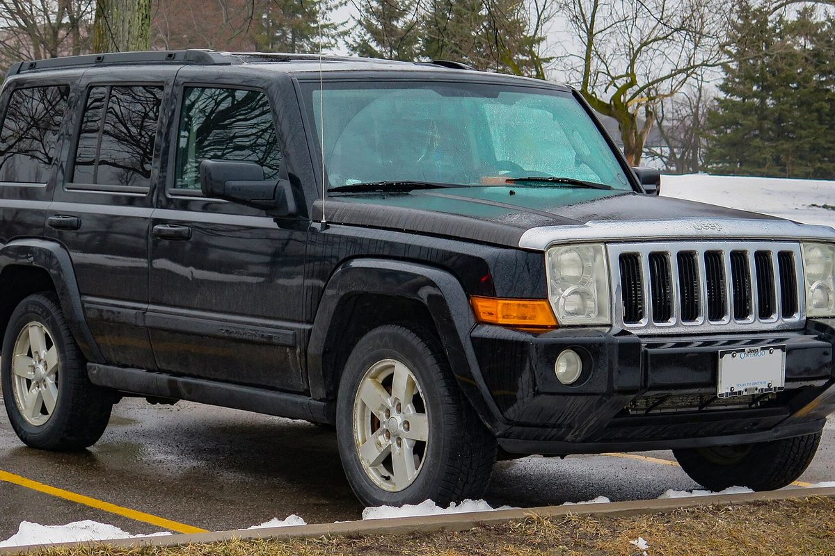 A black Jeep Commander Sport with silver alloy wheels and a chrome grille, parked on a wet lot with patches of snow