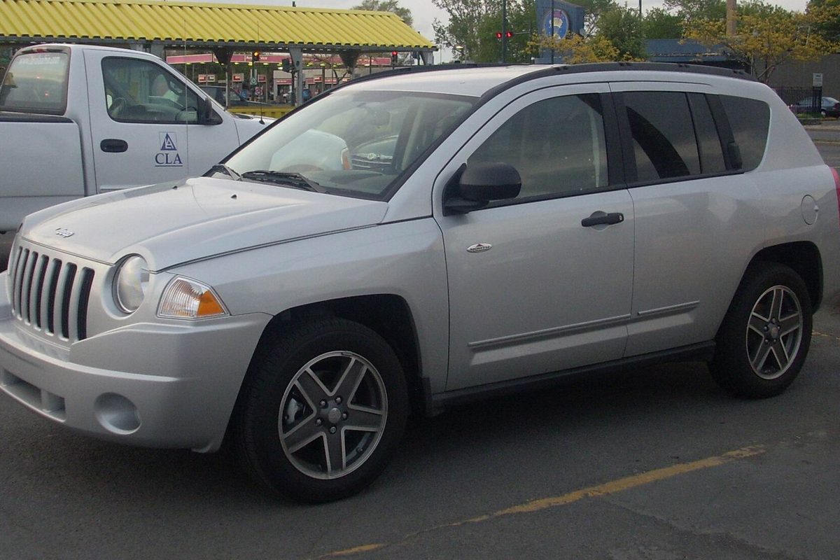 A silver first-generation Jeep Compass with alloy wheels, parked in a lot beside a white pickup truck