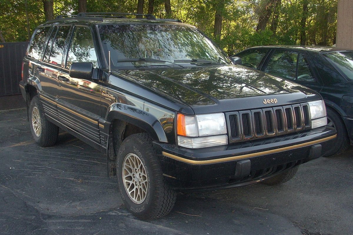 A black first-generation Jeep Grand Cherokee Limited with gold lower trim and lattice alloy wheels, parked on wet asphalt under trees