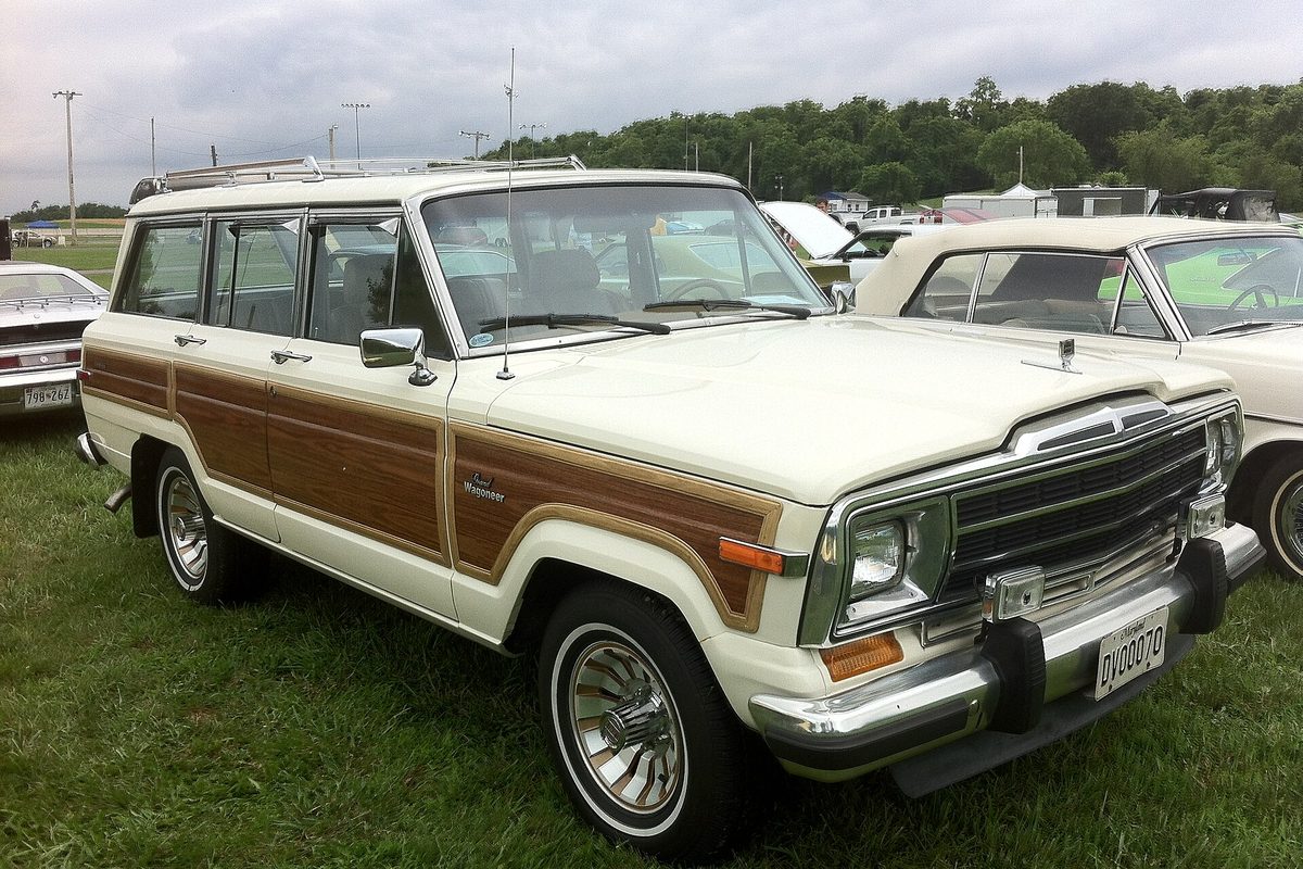 A white 1986 Jeep Grand Wagoneer with woodgrain side panels and wire wheel covers, parked on grass at a car show