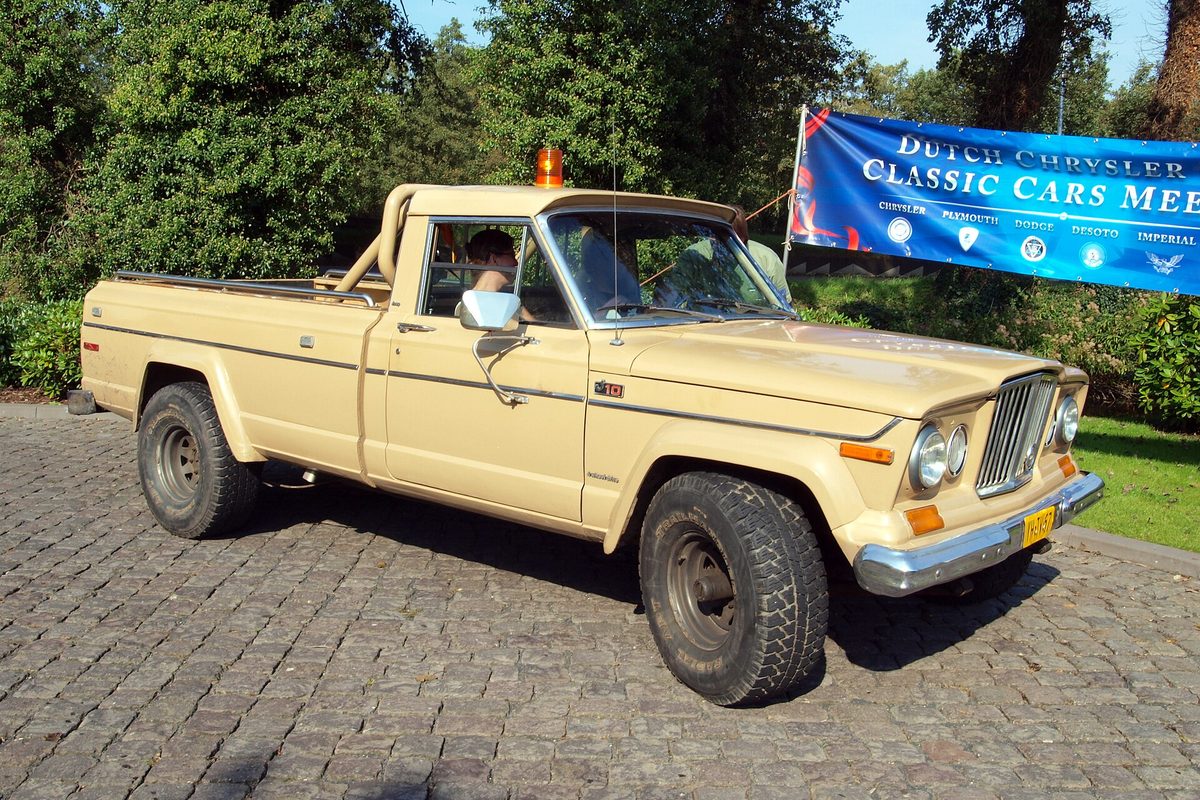 A cream-colored Jeep J10 long-bed pickup with a roll bar, chrome front bumper and black steel wheels, parked on cobblestones at a car show