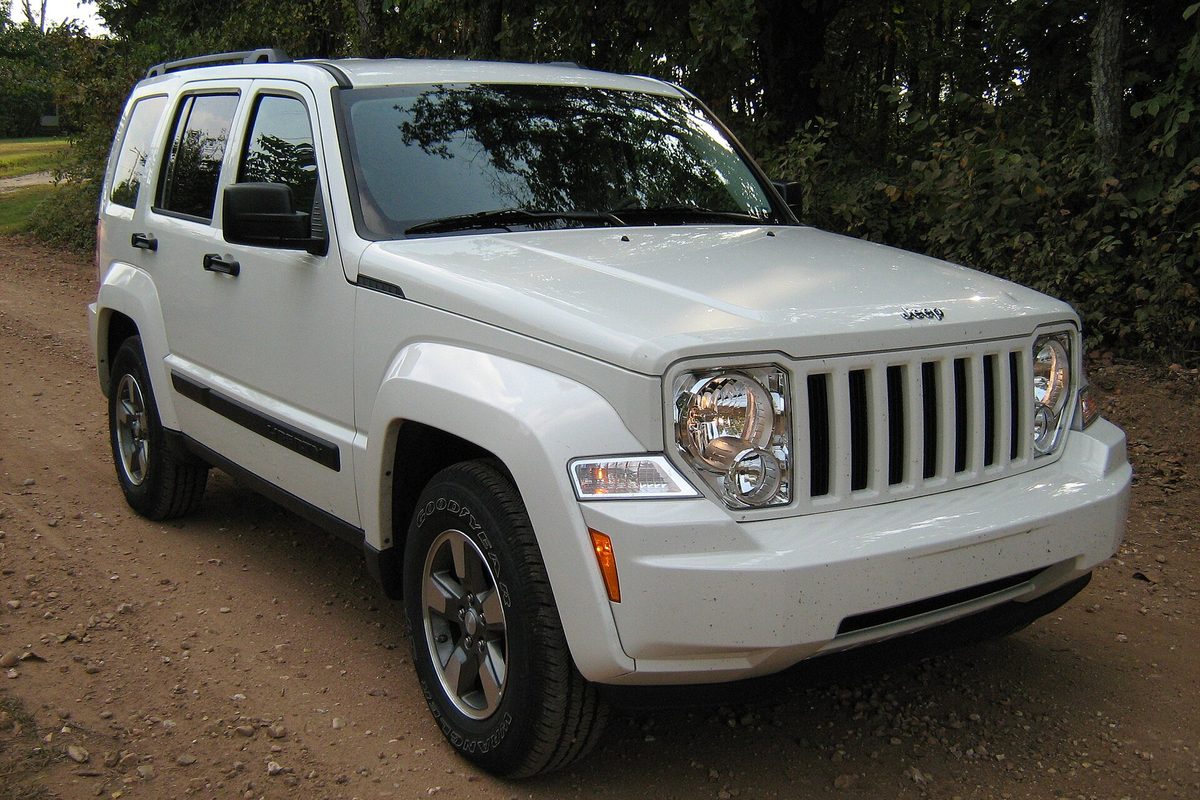 A white second-generation Jeep Liberty with a roof rack and round headlights, parked on a dirt road by a wooded field