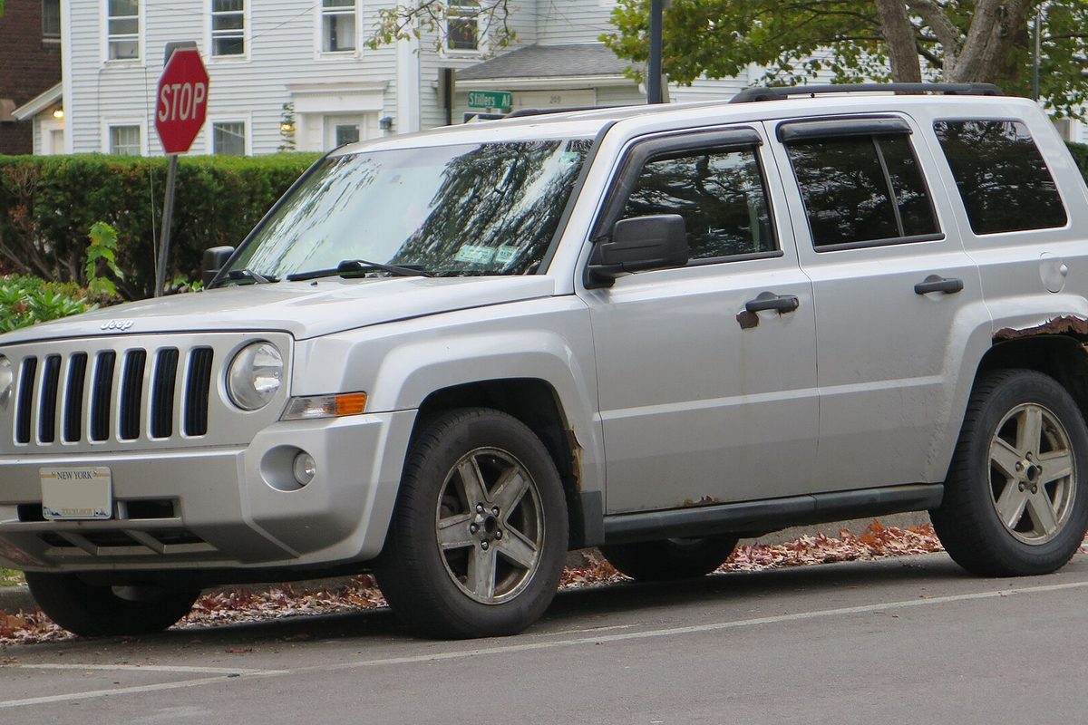 A silver 2010 Jeep Patriot with rust along the rear wheel arch and lower doors, parked at a curb beside a stop sign