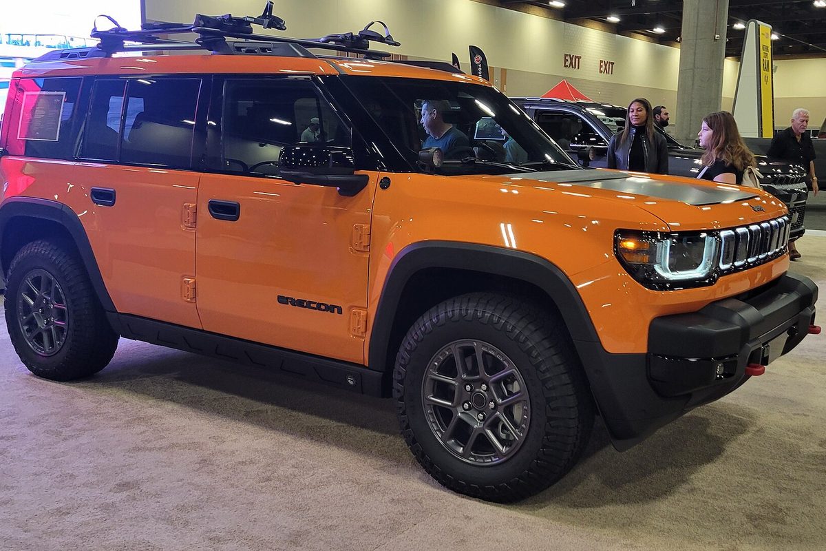 An orange Jeep Recon Moab electric SUV with black fender flares, off-road tires and a roof rack, on display at an indoor auto show