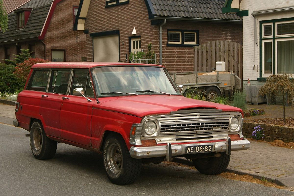 A faded red early full-size Jeep Wagoneer with a chrome grille and bumper, parked on a street of brick houses