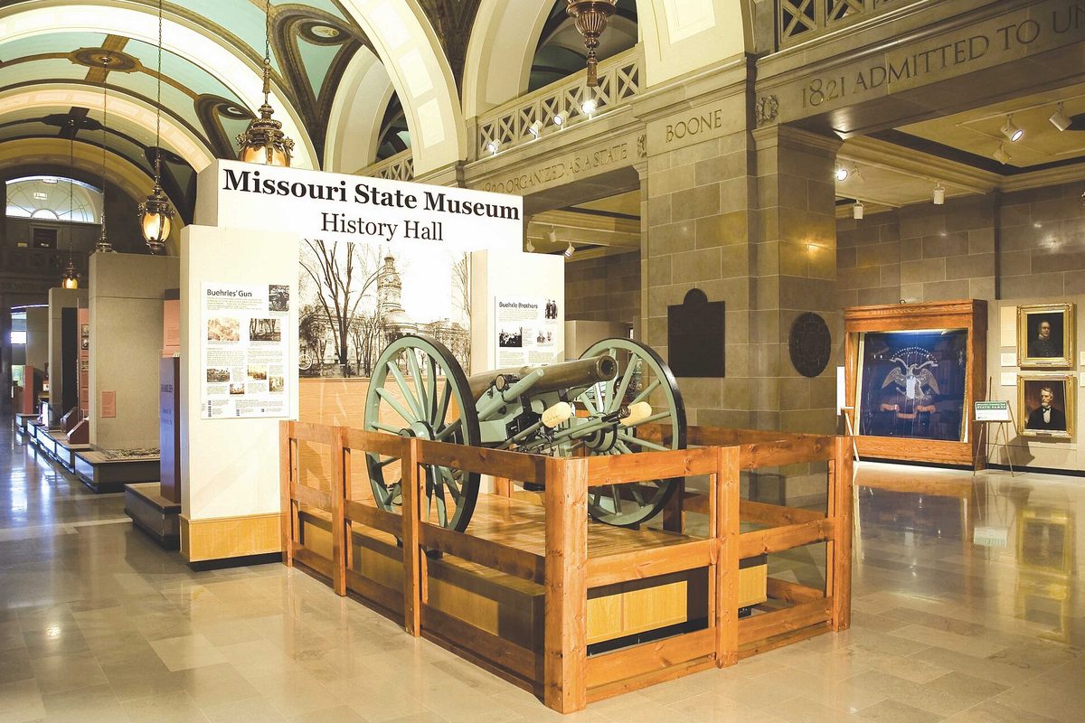 Inside the Missouri State Museum's History Hall, a field cannon behind a wooden rail beneath a painted, arched ceiling with hanging lanterns