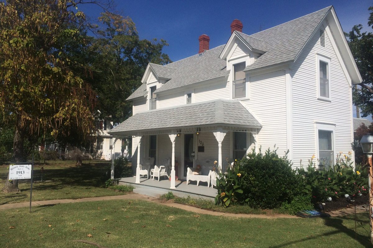 The white two-story B.W. McLean House in Jenks, a frame house with a gray roof, red chimneys and a front porch with white wicker chairs, and a small sign on a post in the yard