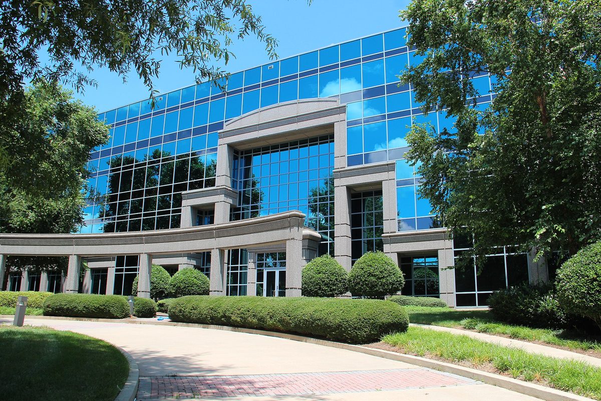 The glass office building that houses Johns Creek City Hall, with a curved stone colonnade, rounded hedges and shade trees on a sunny day
