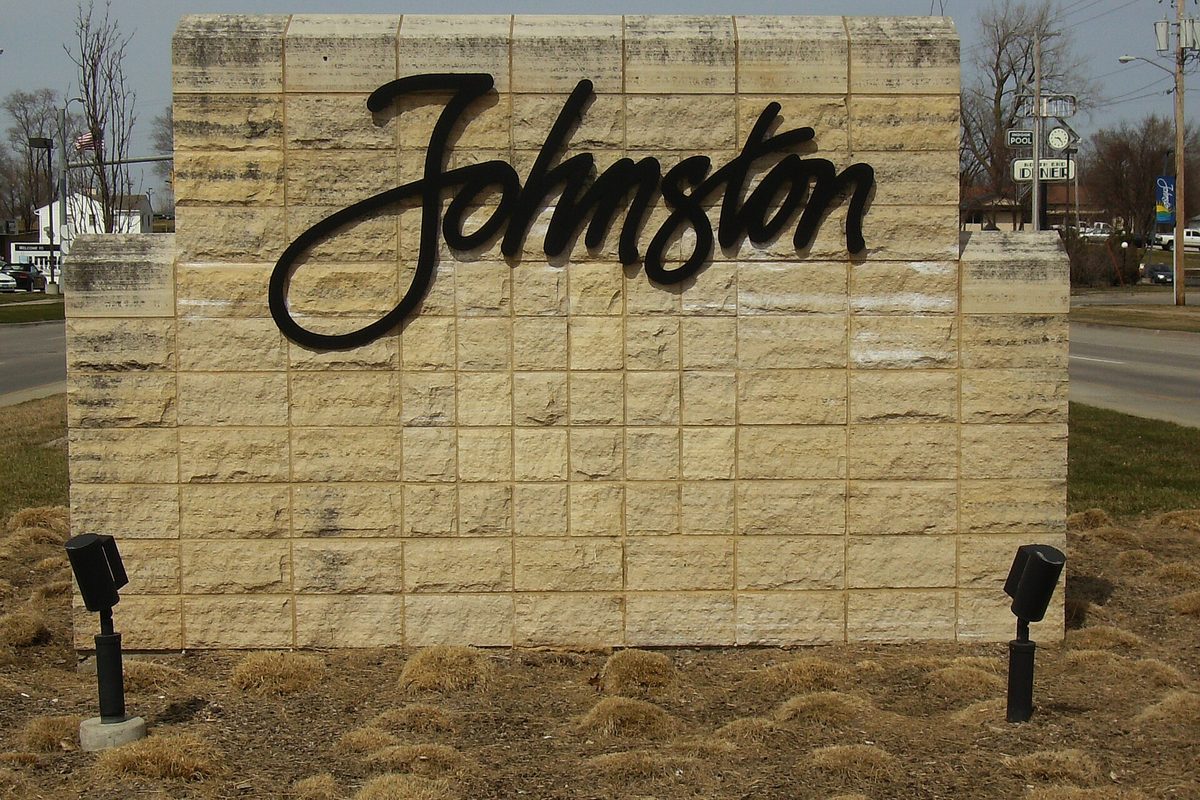A tan stone block monument sign with Johnston written in black script, two small spotlights in the dry grass in front of it