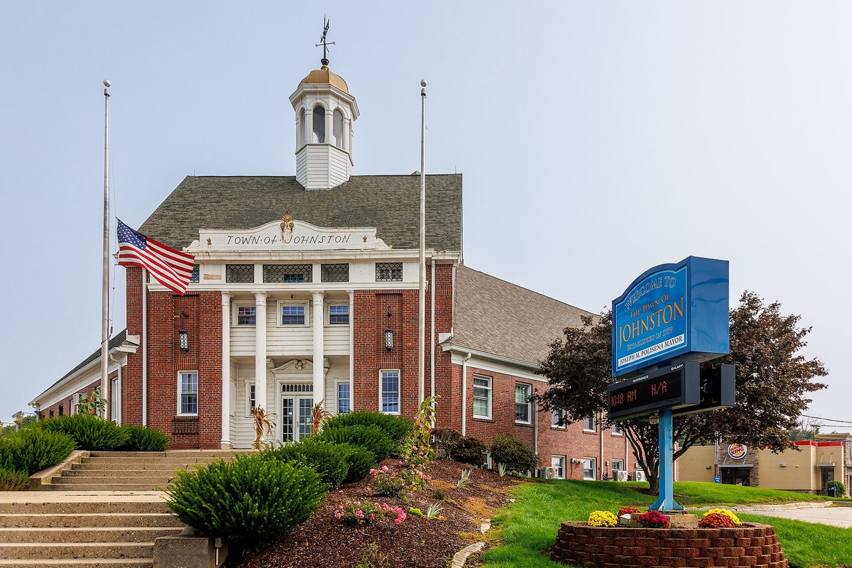 The red brick Johnston Town Hall with white columns, a white cupola and a flag at half staff, beside a blue Johnston welcome sign and a flower bed