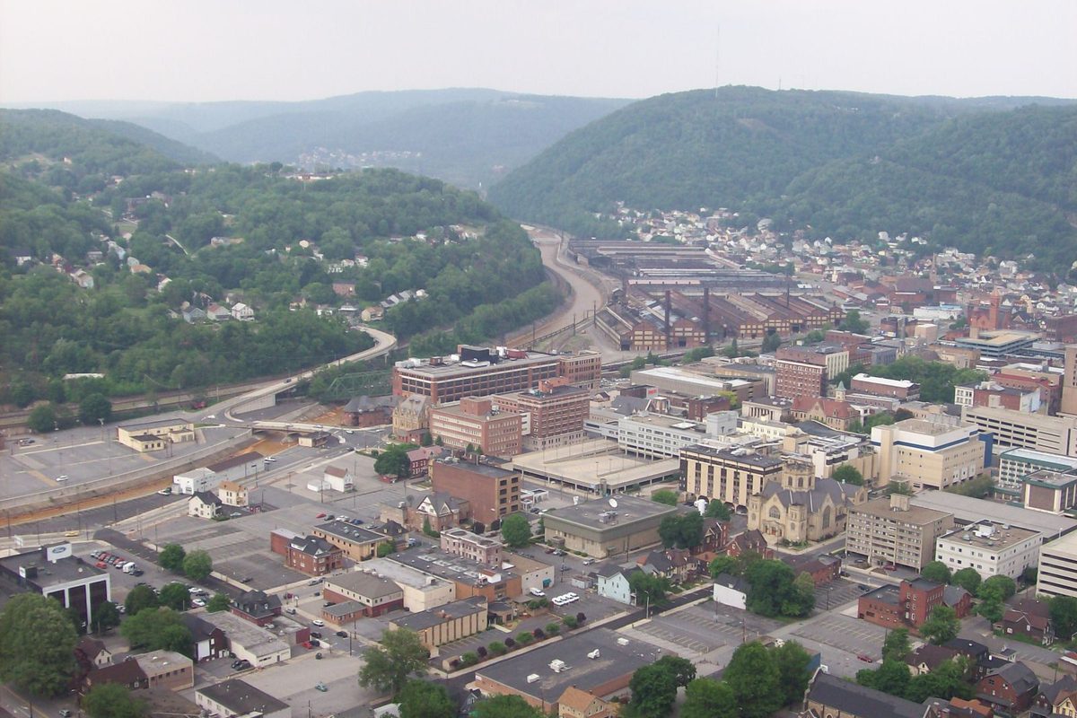 Downtown Johnstown seen from above, brick blocks and church towers in the foreground and long mill buildings and rail lines along the valley floor between green hills