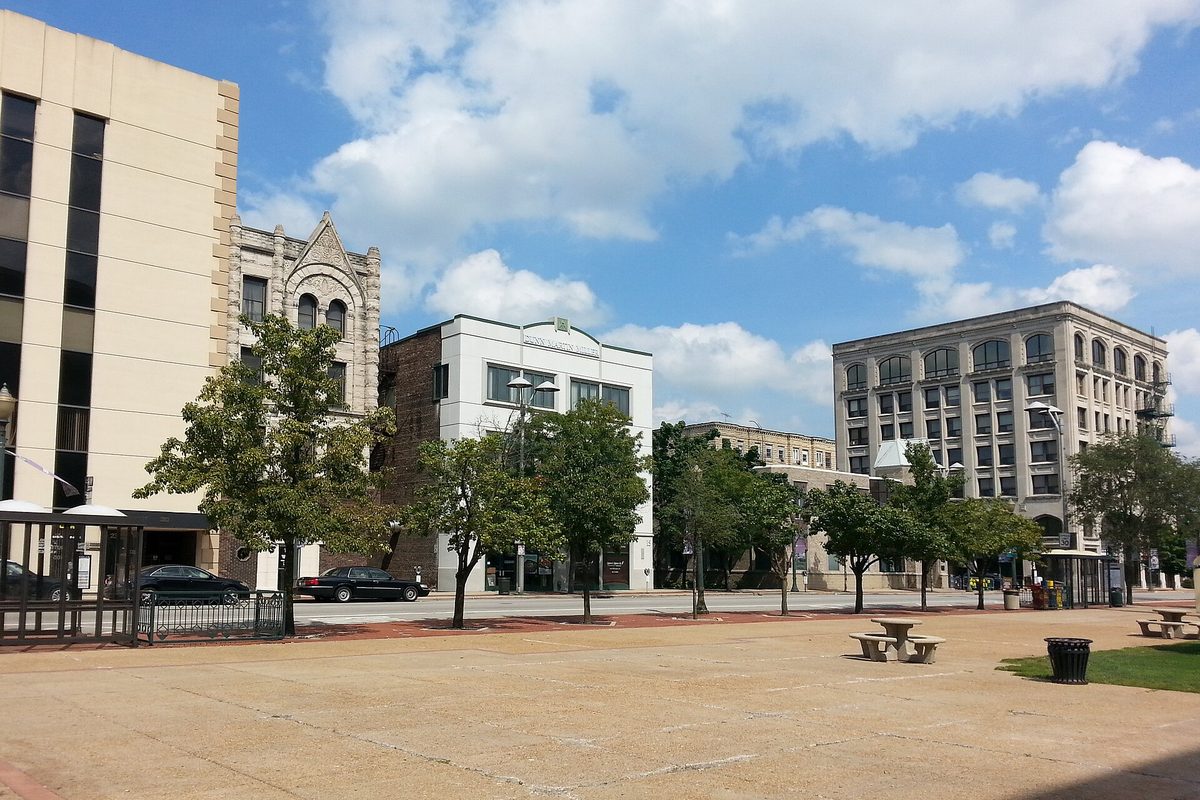 A wide open plaza in downtown Joliet with picnic tables, young trees and a row of older stone and brick office buildings under a blue sky with white clouds