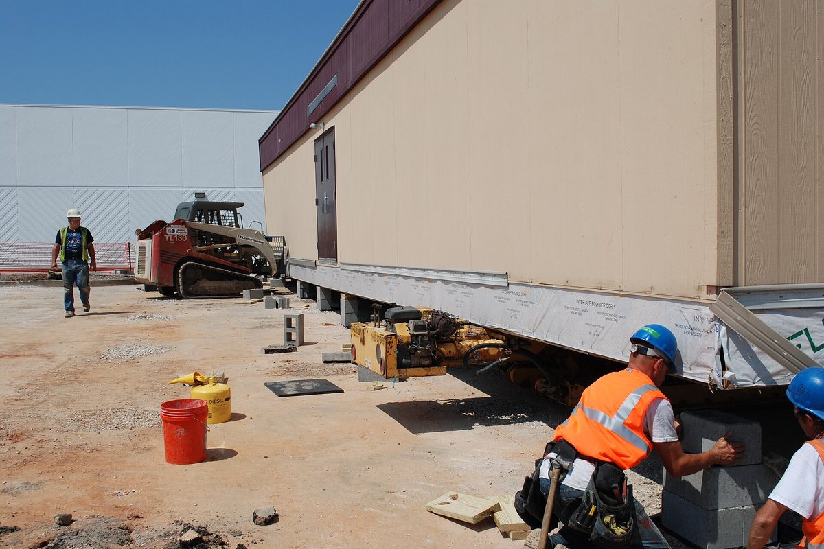 Workers in hard hats and safety vests set blocks under a modular classroom building at the former Shopko site in Joplin, with a skid steer behind them