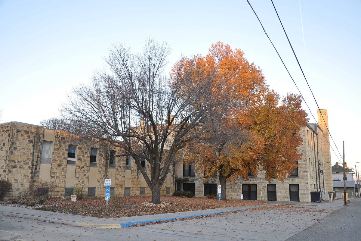 The tan stone First Presbyterian Church of Junction City on a street corner, behind a bare tree and a tree in orange autumn leaves