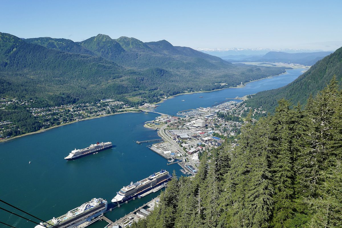 Downtown Juneau seen from a forested mountainside, with three large cruise ships docked along a blue channel and green mountains beyond