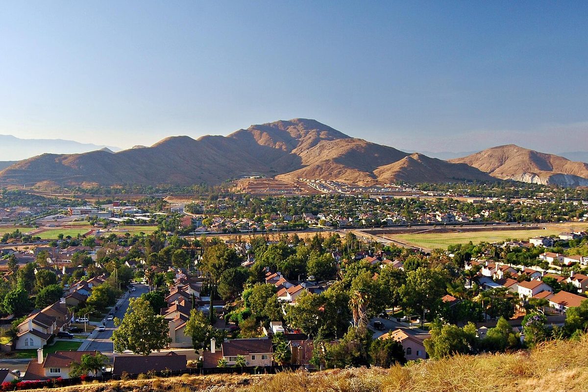 The brown slopes of Mount Jurupa rising above tree-lined streets and rooftops in Jurupa Valley, seen from a dry grassy hillside