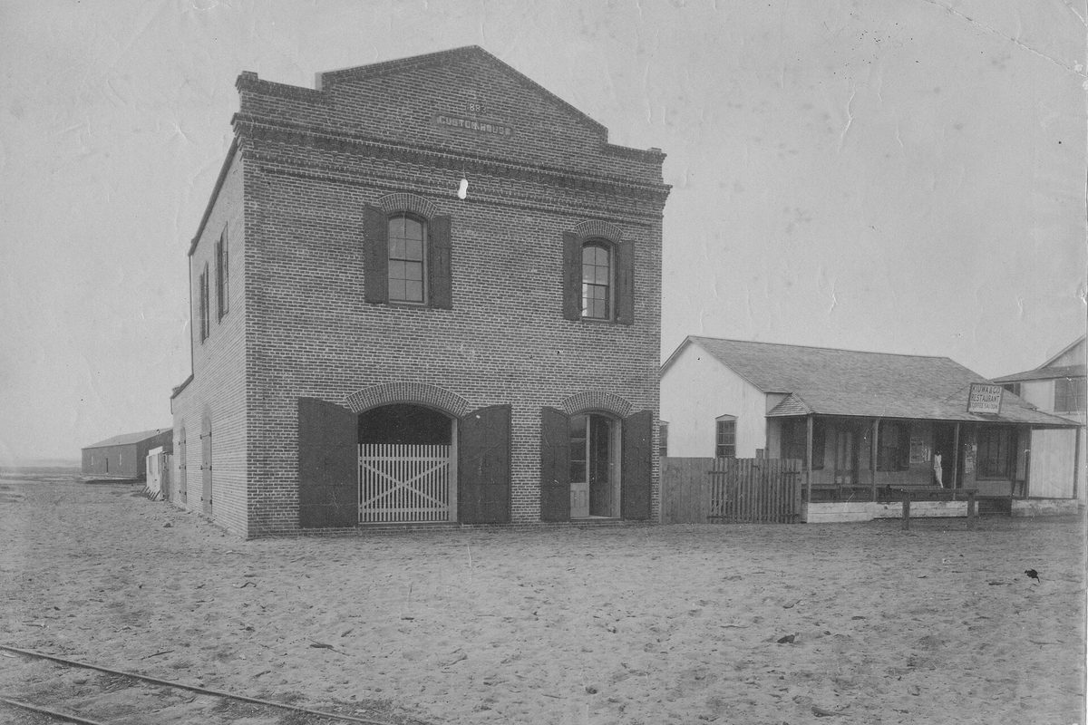 Old sepia photograph of the two-story brick customs house at Kahului, Maui, with arched doorways, a wooden house beside it and rail tracks in the sandy foreground