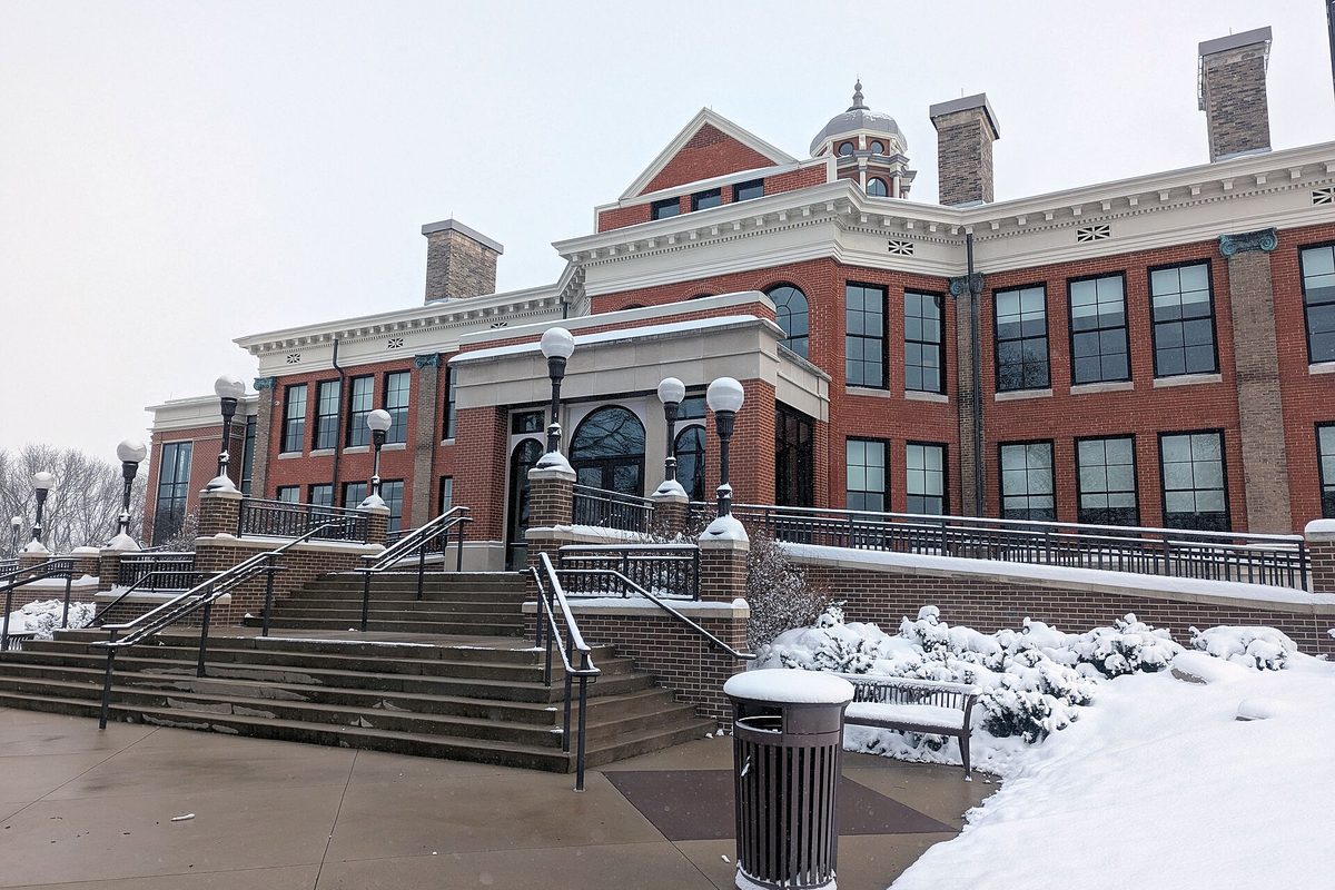 Western Michigan University's Heritage Hall, a red brick building with white trim and a cupola, behind snow-covered steps, railings and shrubs
