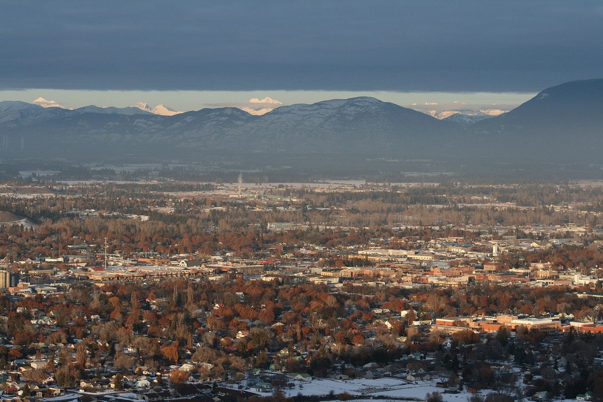 Kalispell at dusk from a hilltop, rows of houses among rust-colored autumn trees, with snow on the ground and snowy mountain peaks under dark clouds beyond