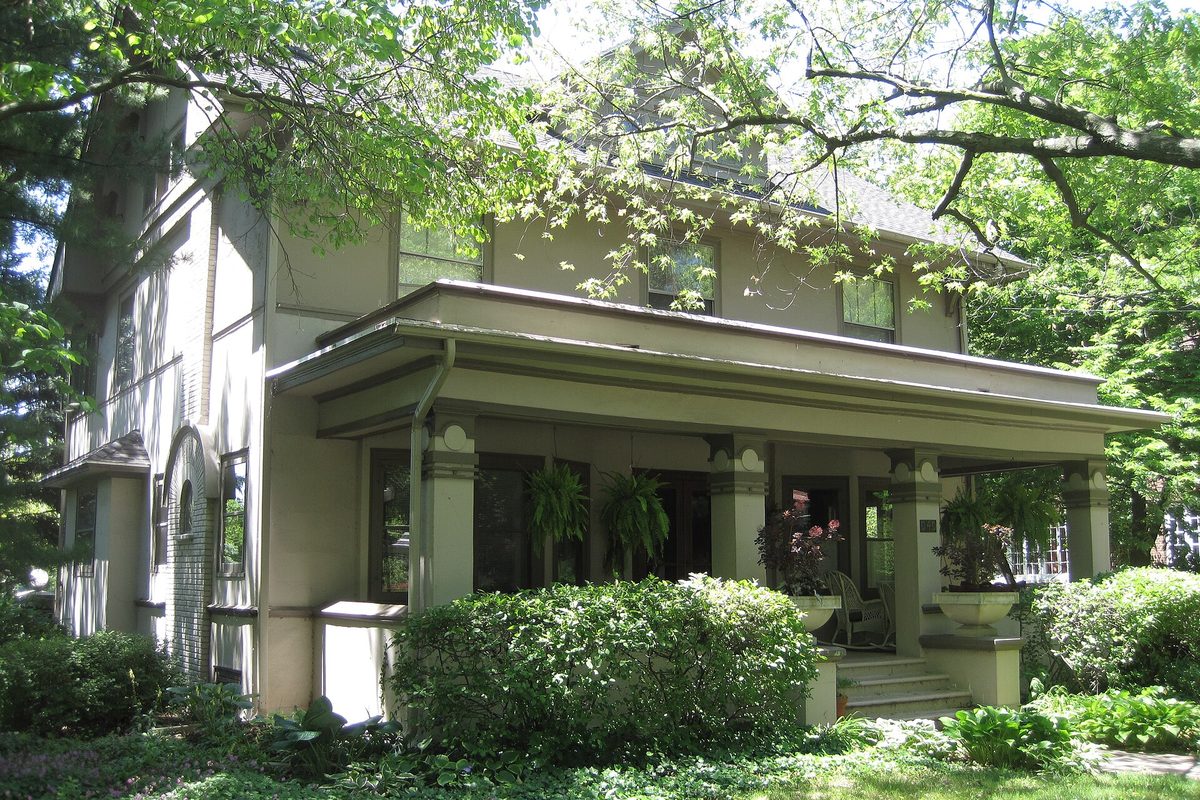 The two-story stucco Wilhemine Mang House with a broad covered front porch and potted ferns, shaded by trees in Kankakee's Riverview Historic District