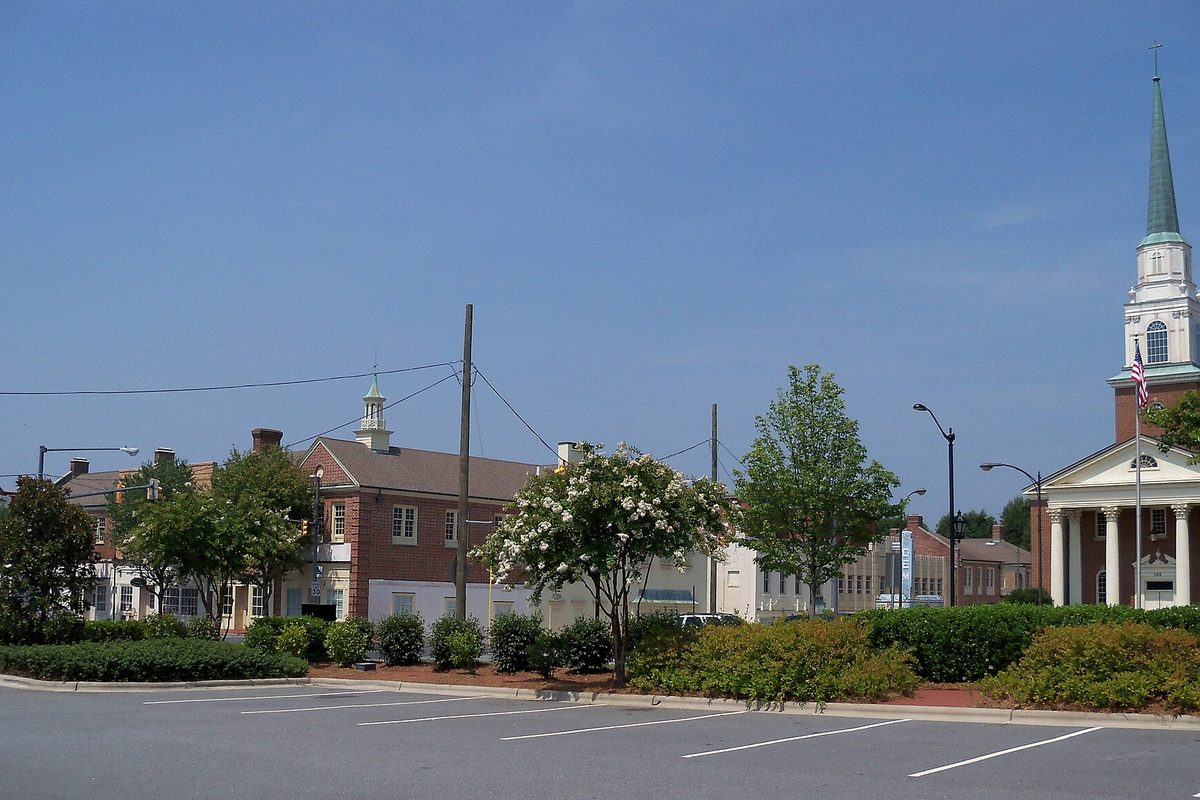 Red brick buildings and a white church steeple beside an empty parking lot in Kannapolis, with a flowering white crape myrtle under a blue sky