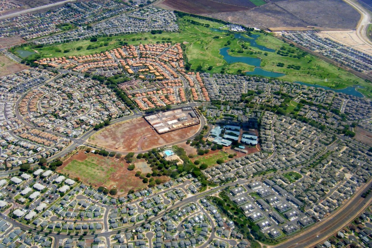 Aerial view of Kapolei's tract-home neighborhoods around a large bare dirt lot, with a golf course and blue ponds beyond