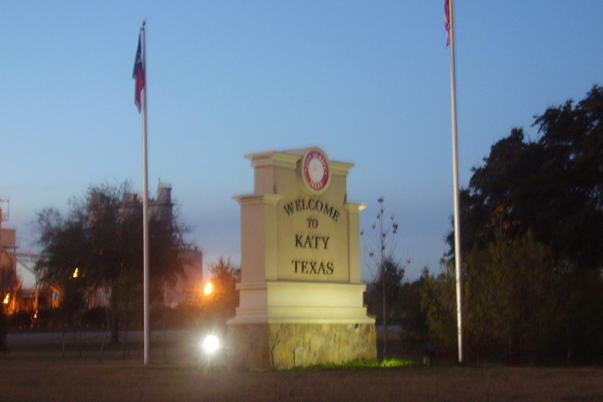 A lit stone monument reading Welcome to Katy, Texas at dusk, between two flagpoles with trees and plant lights behind